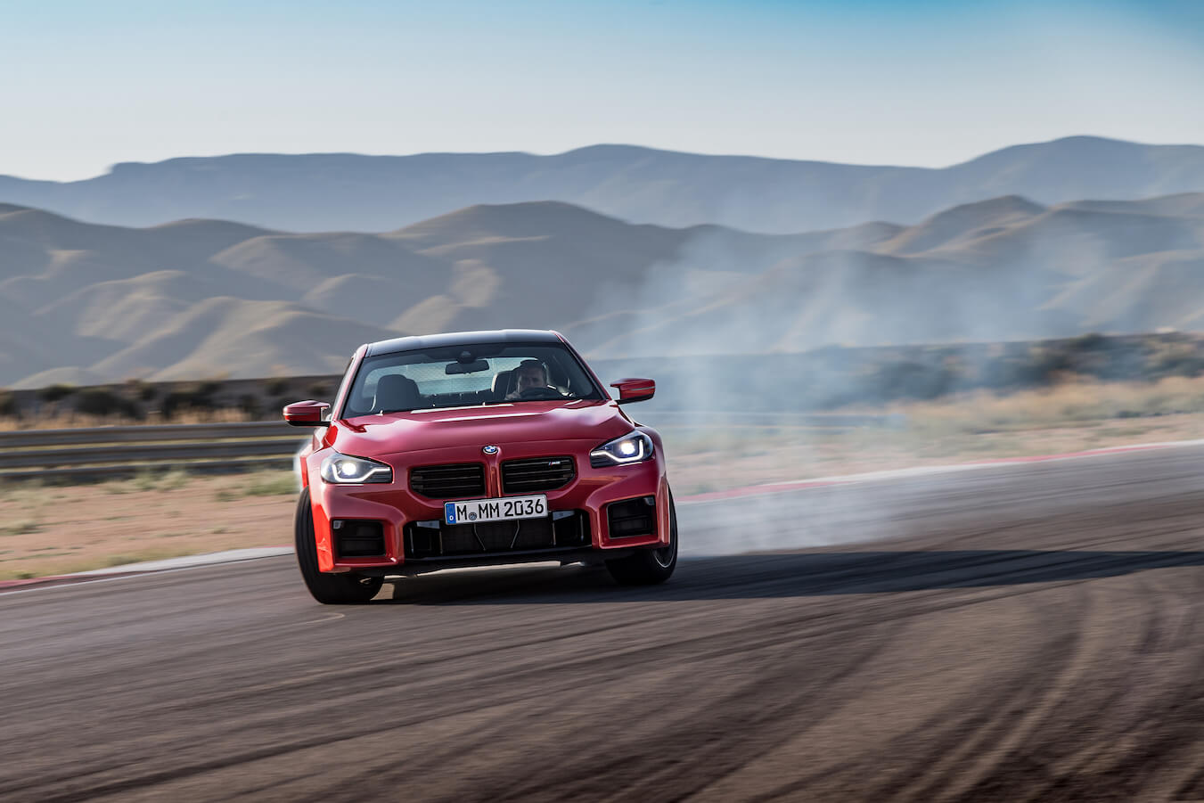A red 2024 BMW M2 driving down a racetrack during the middle of the day.