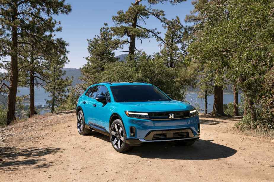A bright blue 2024 Honda Prologue EV parked in right front angle on a dry dirt hill with spruce trees peppered in the background