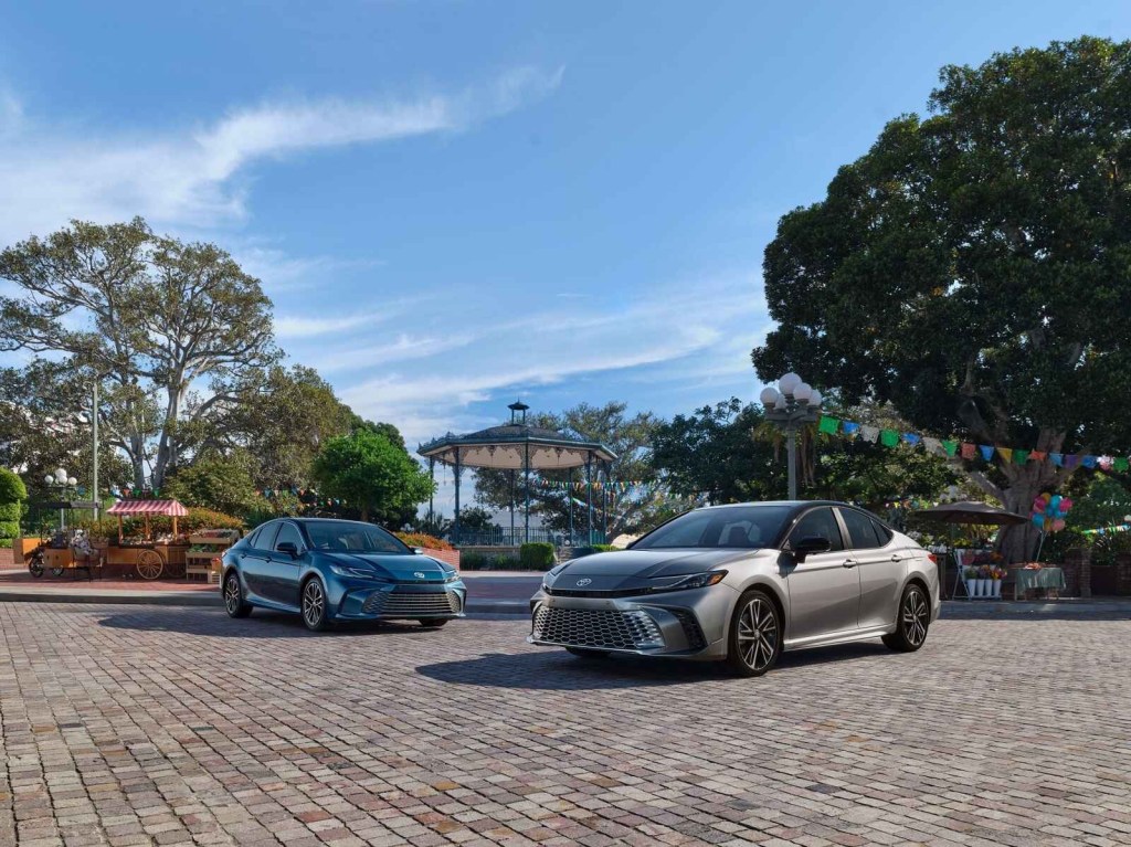 Two 2025 Toyota Camrys, one blue in the background and grey in the mid ground, are parked at angles on a paver stone area