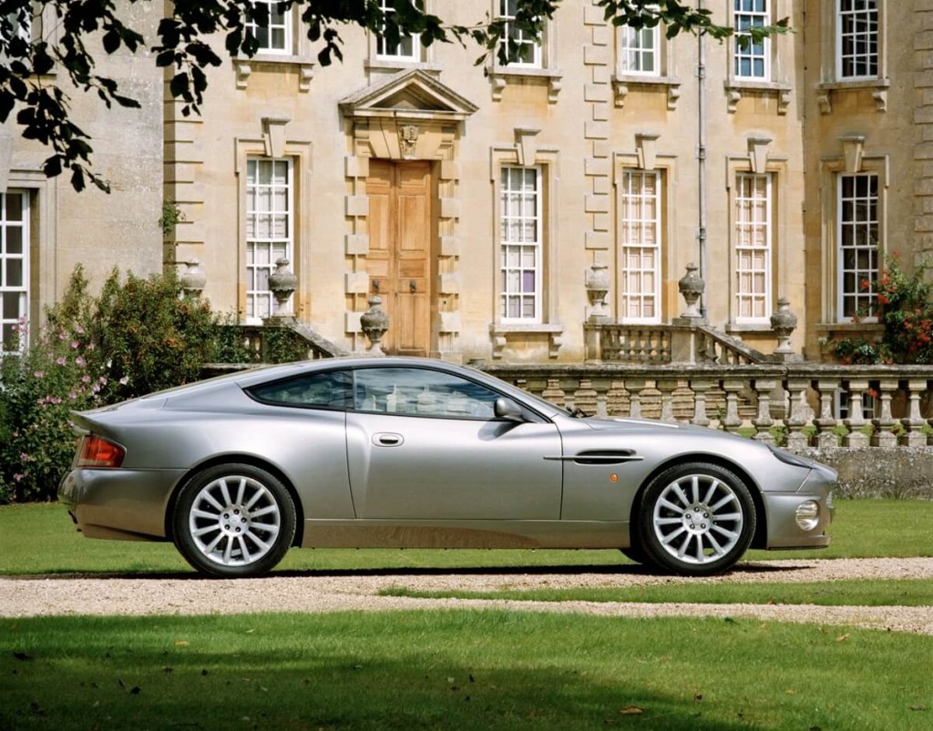 A silver Aston Martin V12 Vanquish shows off its lines, which are chunkier than the Vantage, as it sits by a manor. 
