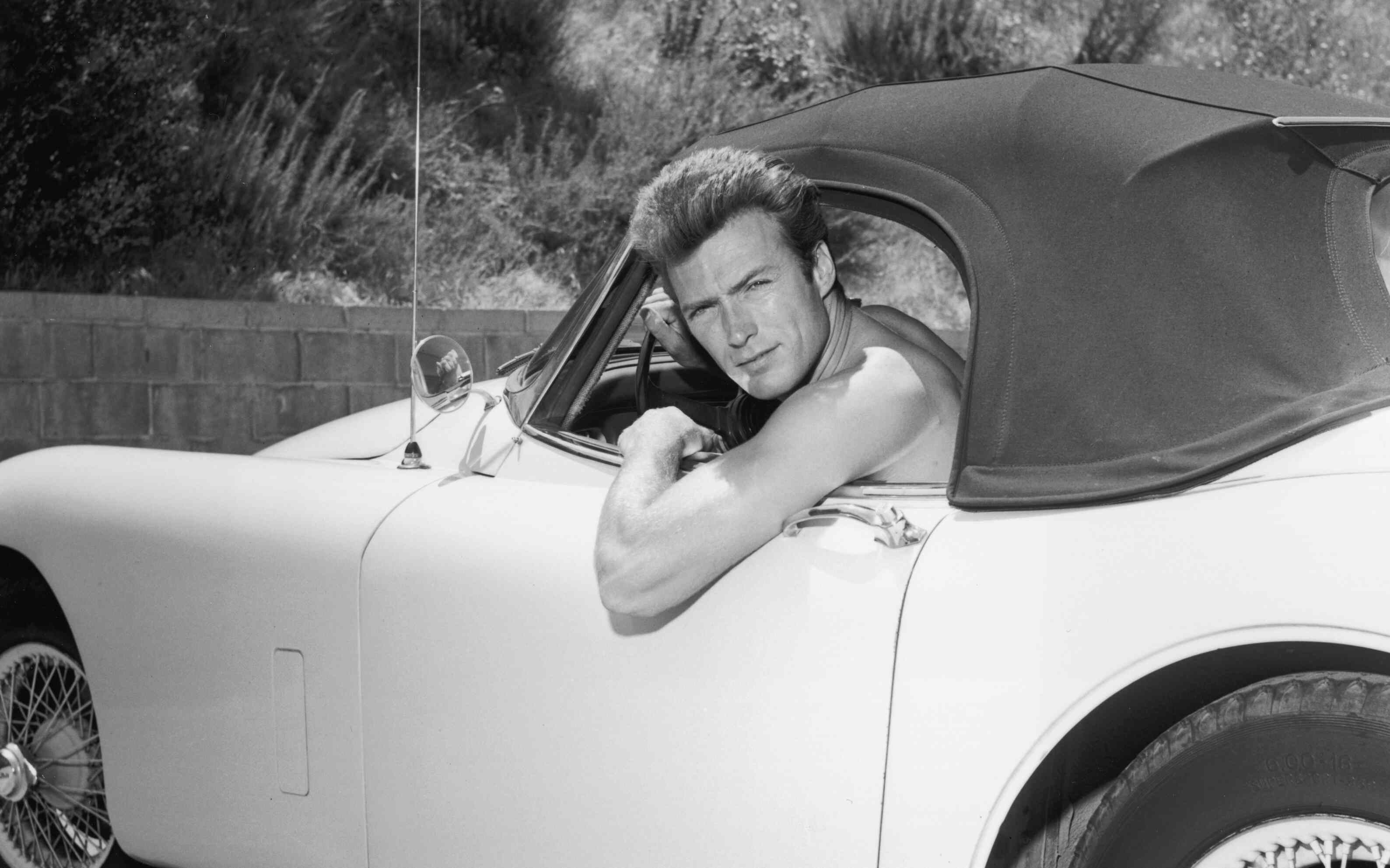 A shirtless Clint Eastwood leans out the window of a Jaguar convertible (ca. 1965)