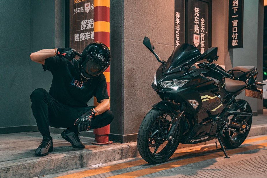 Motorcyclist crouches next to his Kawasaki Ninja while wearing a black motorcycle helmet.