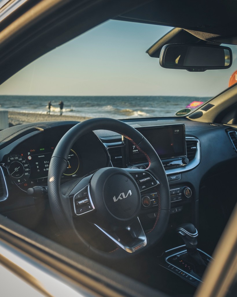 An interior shot of a Kia car with the Kia emblem displayed on the steering wheel with the beach and ocean in the background