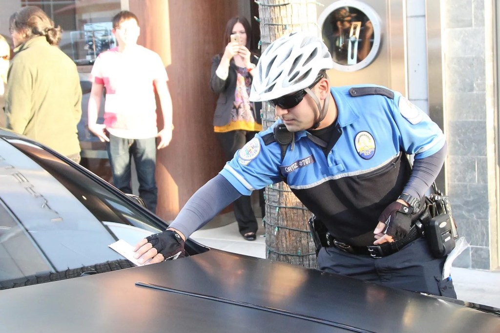 Man in a blue uniform and bicycle helmet leaves a parking ticket on a car's windshield.