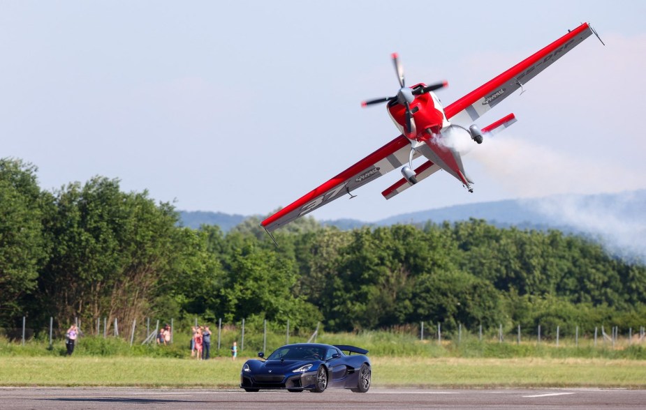 A Rimac Nevera electric supercar races down a runway while a prop plane flies overhead.