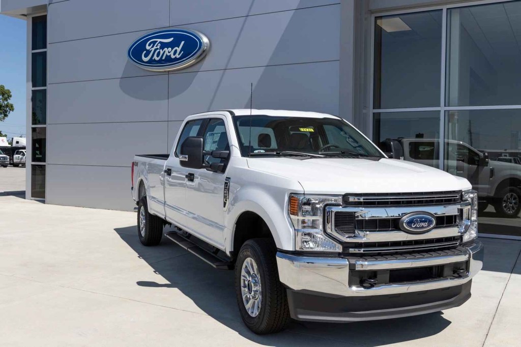 A white Ford truck is parked in right front facing angle in front of a Ford dealership