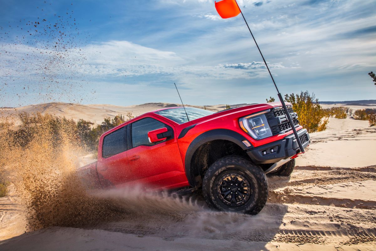 A 2023 Ford F-150 Raptor R full-size pickup truck model driving in sand off-road with a red flag attached