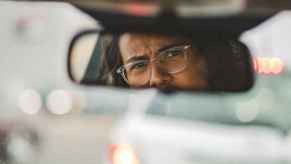 A man with glasses expresses questioning emotions in his review mirror possibly at a tailgating car
