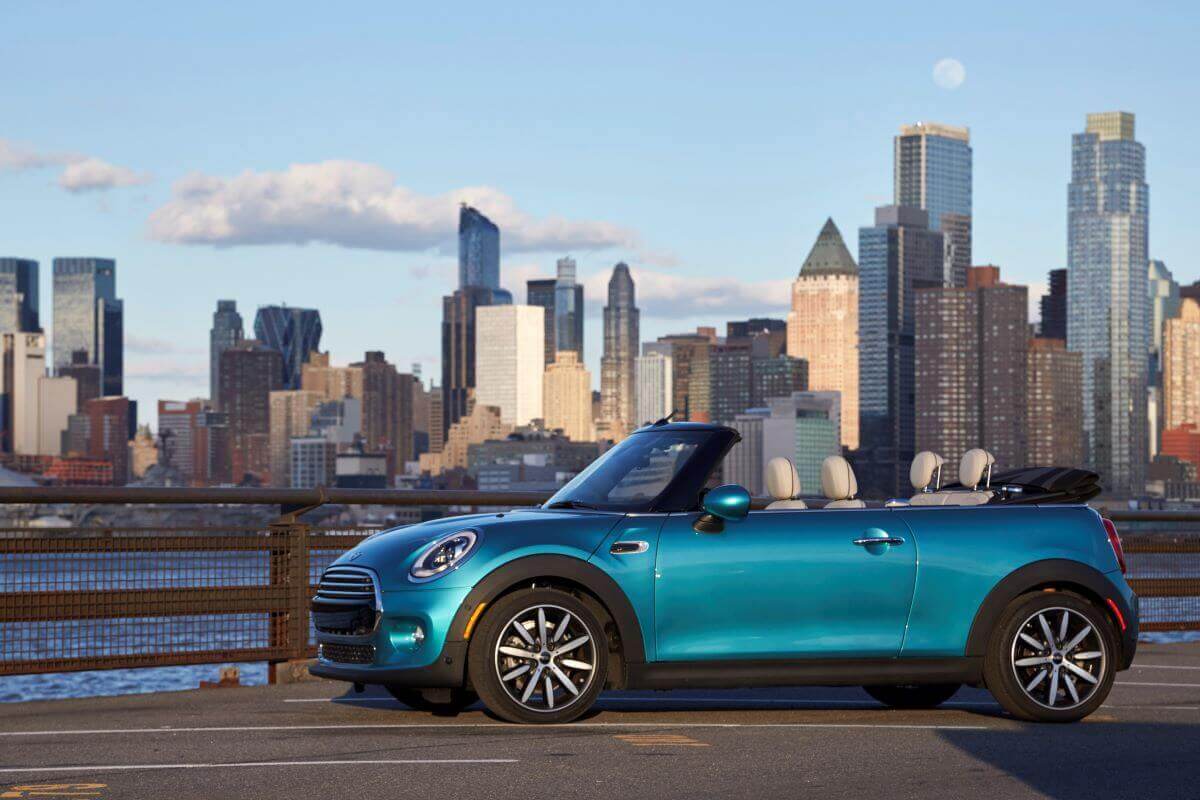 A Mini Cooper Convertible compact car model with its tops down parked near a river and city skyline background