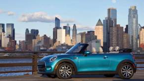 A Mini Cooper Convertible compact car model with its tops down parked near a river and city skyline background