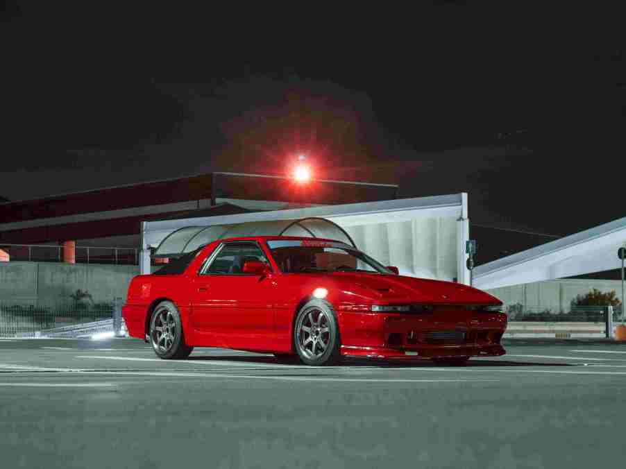 A red Toyota Mk3 Supra is shown in a parking lot at night.