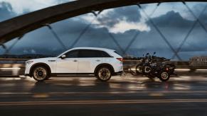 An exterior side profile shot of a 2024 Mazda CX-90 PHEV full-size SUV towing a pair of bikes across a bridge