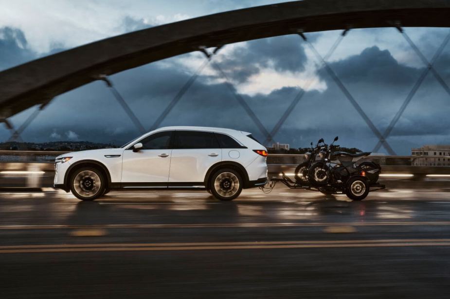 An exterior side profile shot of a 2024 Mazda CX-90 PHEV full-size SUV towing a pair of bikes across a bridge