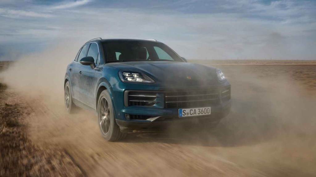 A 2023 Porsche Cayenne SUV is driving to the right of the frame on a dusty road
