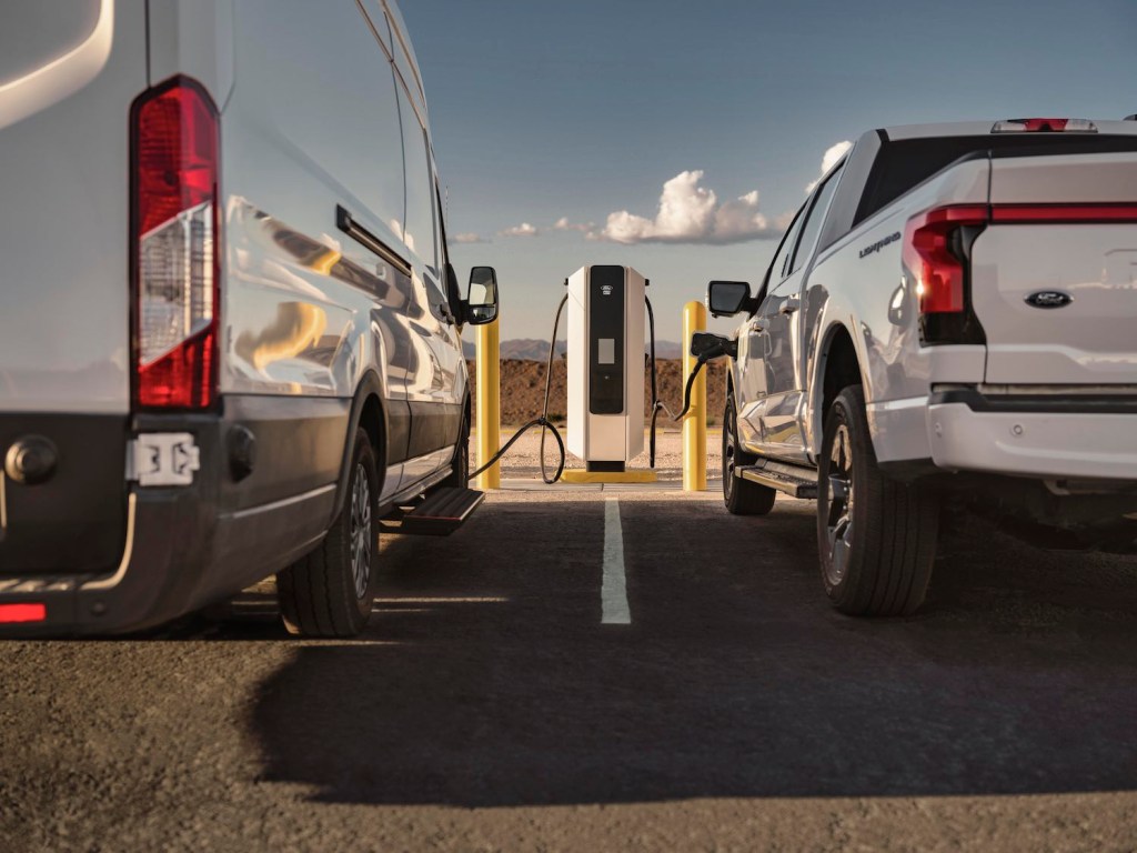 The rear bumpers of an electric F-150 and Ford Transit van both plugged into a charger.