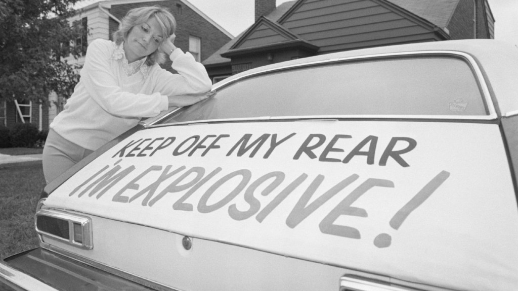 Woman leaning on a Ford Pinto with a warning of the dangers of hitting this  car's rear.