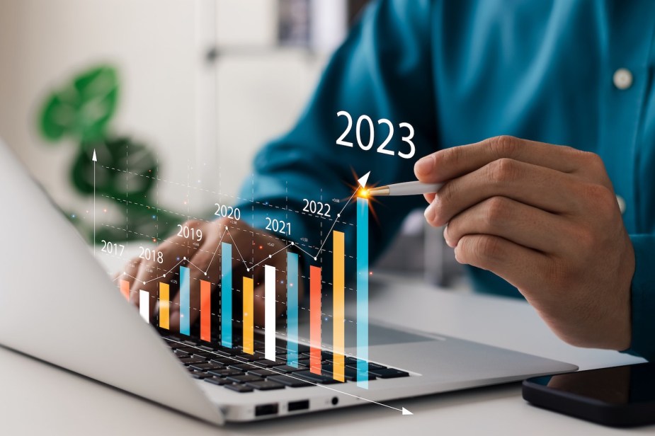 Graphic of a hand pointing at a yearly prediction going up in 2023 above a laptop.