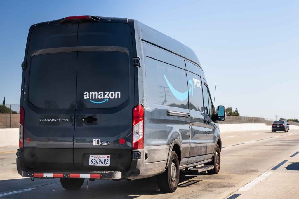 A blue Amazon Ford Transit delivery van is shown on the highway in right rear view.