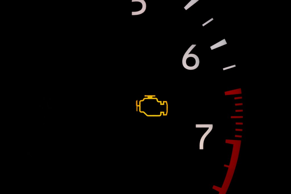 Check engine light illuminated in yellow is shown close up on a tachometer with white numerals