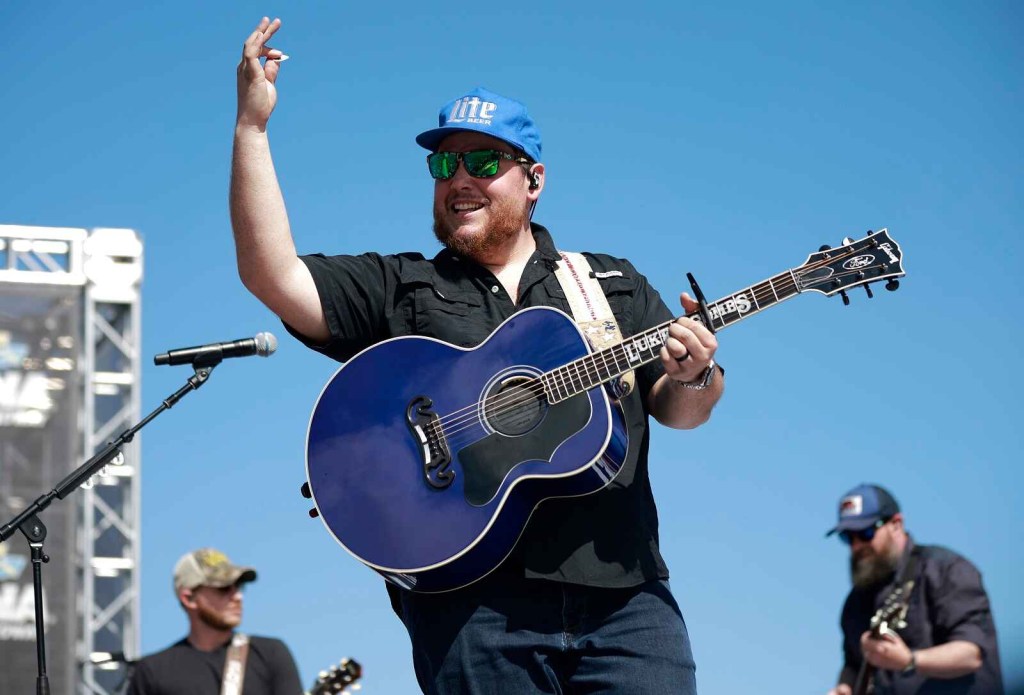 Luke Combs shown waving from an outdoor stage with his blue guitar