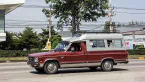 A burgundy colored Mazda B2200 compact pickup truck with an aluminum bed cap drives on the road in left profile view
