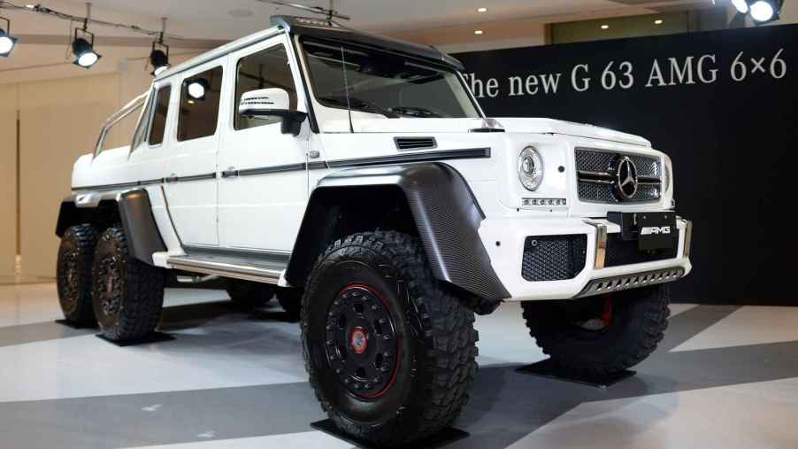 A white Mercedes-Benz G63 6x6 pickup is shown parked at a right front angle on a showroom floor