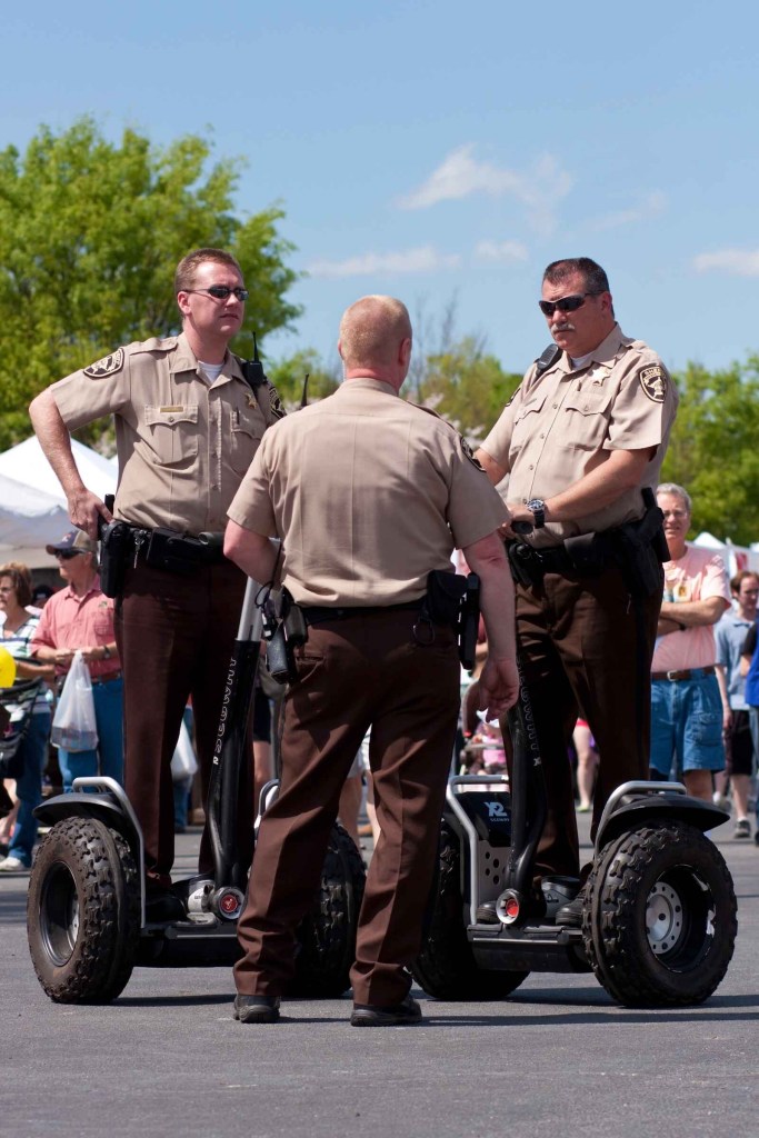 A group of police officers in brown uniforms face each other two are riding Segways