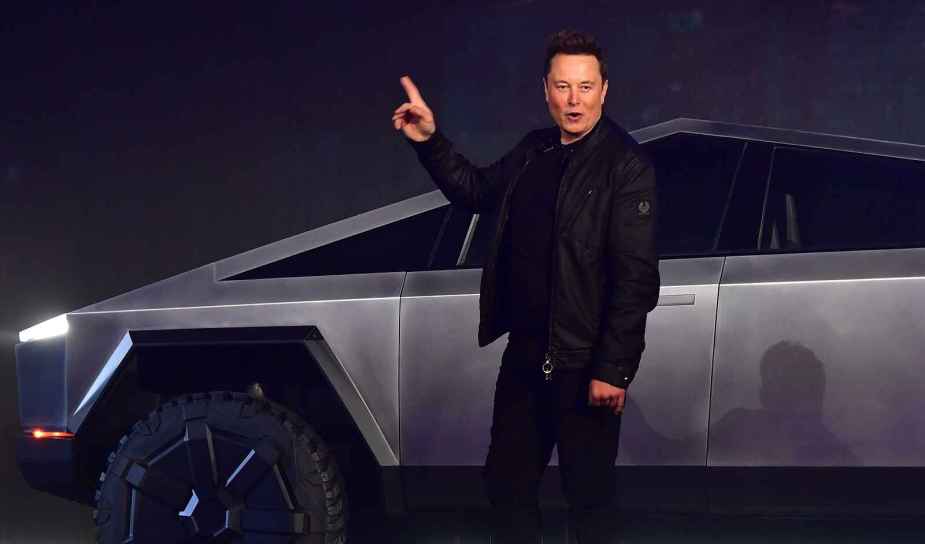 Elon Musk stands in front of a Tesla Cybertruck with a raised right arm pointing index finger