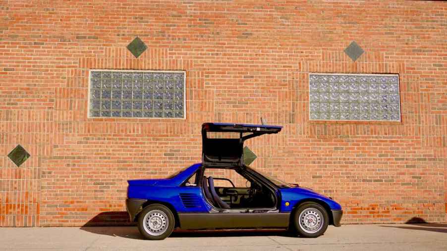 Blue Autozam AZ-1 sports car by Mazda with its gull-wing door open, a brick wall visible in the background