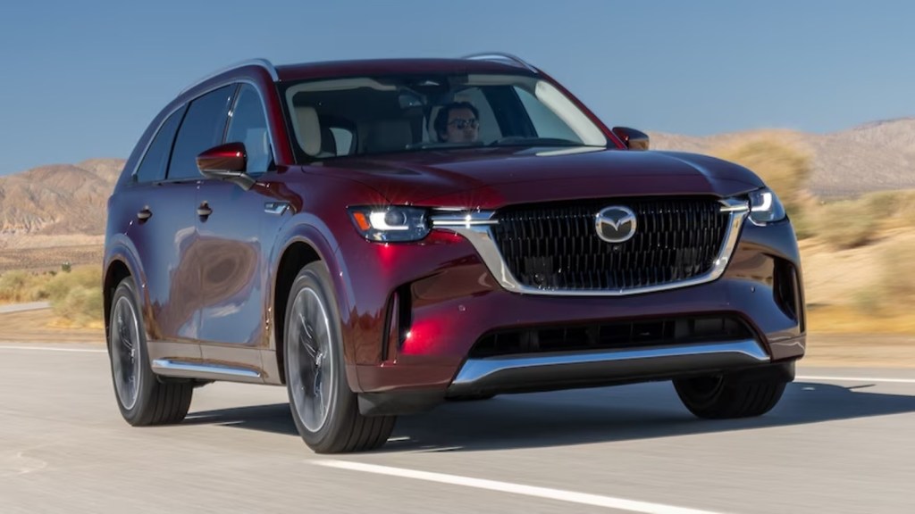 Red 2024 Mazda CX-90 driving on a road. This three-row midsize SUV brings a lot to the table.