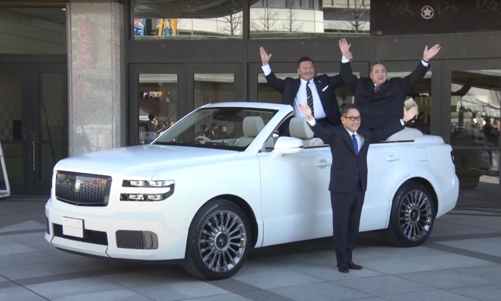 Two sumo wrestlers and Akio Toyoda pose next to a white Toiyota Crown SUV convertible.