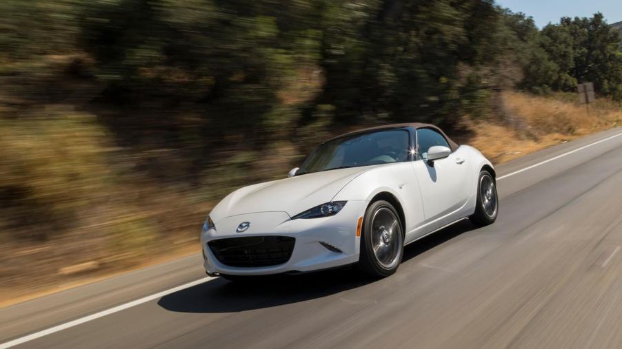 A white Mazda MX-5, a cheaper sports car than a Chevrolet Camaro, drives on an open road.
