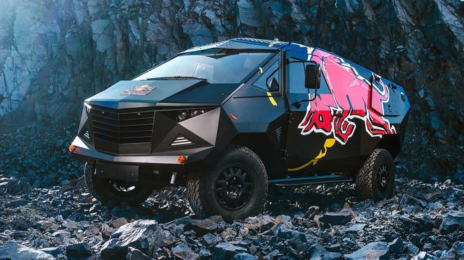 Angular, tactical-style SUV with the red bull logo is parked on rocks.
