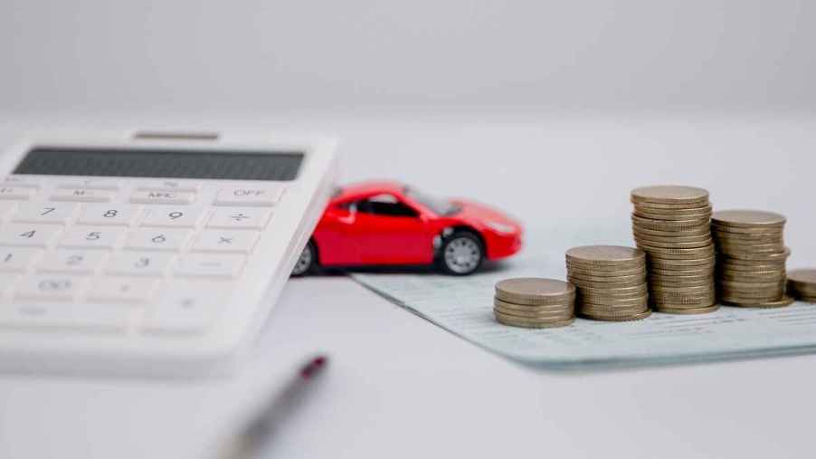 A red toy car is placed on a white surface with a handheld calculator, pen, piece of paper, and stacks of coins