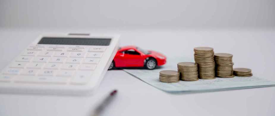 A red toy car is placed on a white surface with a handheld calculator, pen, piece of paper, and stacks of coins