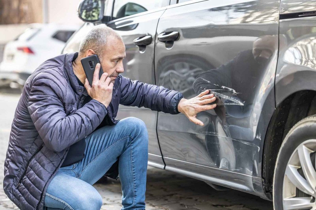 A man kneels next to a scratched black car on his cell phone