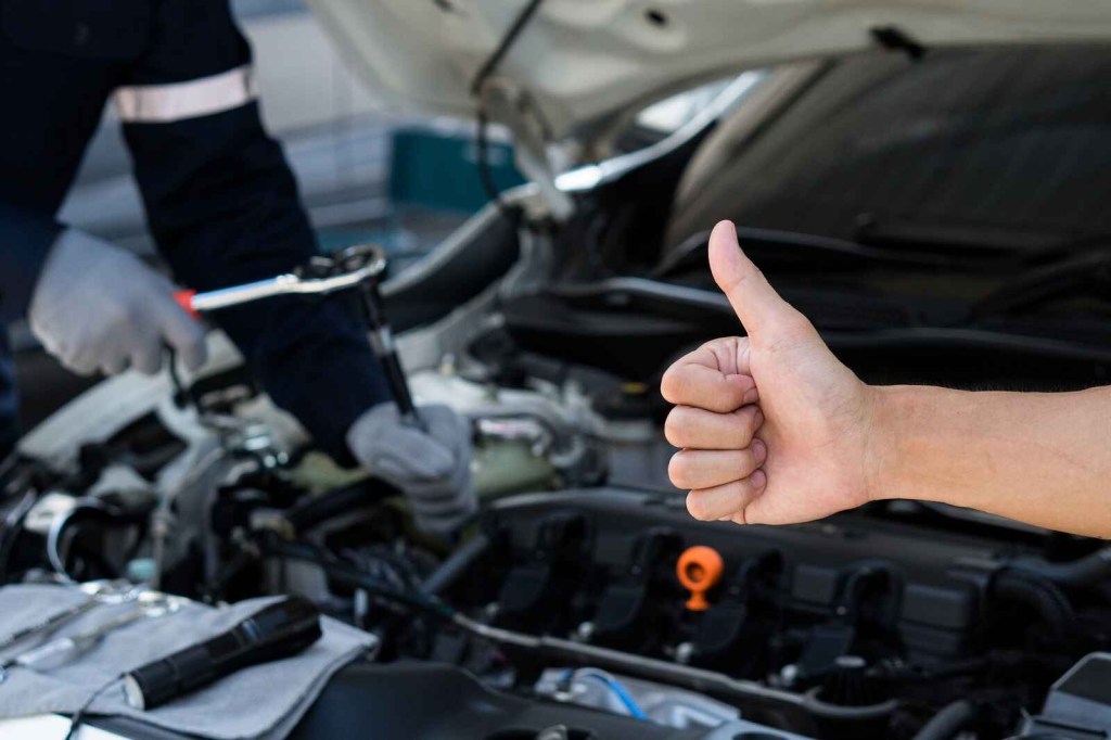 A man's hand gives a mechanic the thumbs-up sign while the mechanic holds a tool and workings in an engine