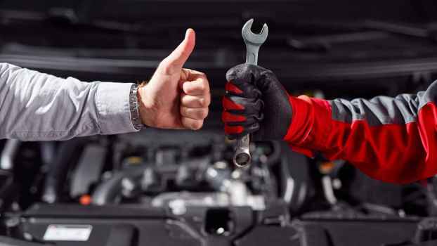 Two forearms with hands, one in a button up shirts and the other a mechanic glove, are shown giving each other thumbs-up in front of an open car engine perhaps agreeing on a car repair estimate