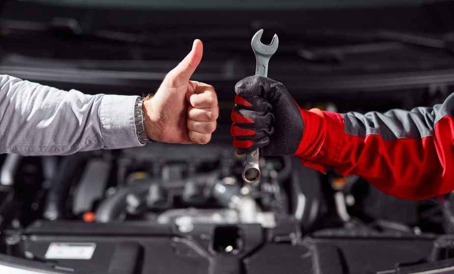 Two forearms with hands, one in a button up shirts and the other a mechanic glove, are shown giving each other thumbs-up in front of an open car engine perhaps agreeing on a car repair estimate