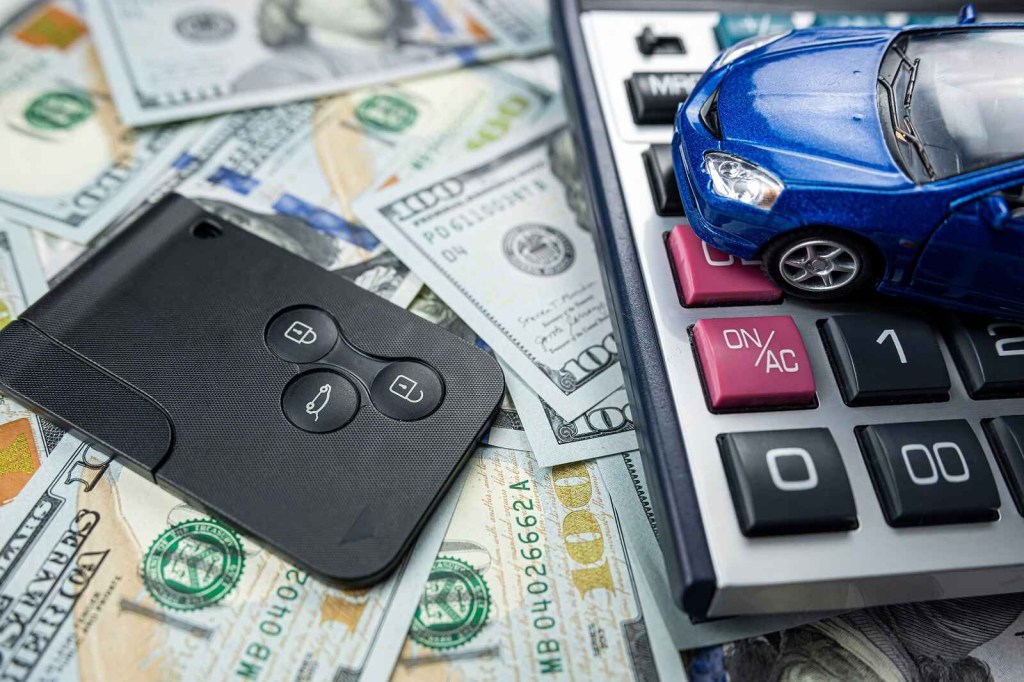 A blue toy car is shown on top of a calculator and spread of U.S. $100 bills with a car key to the left
