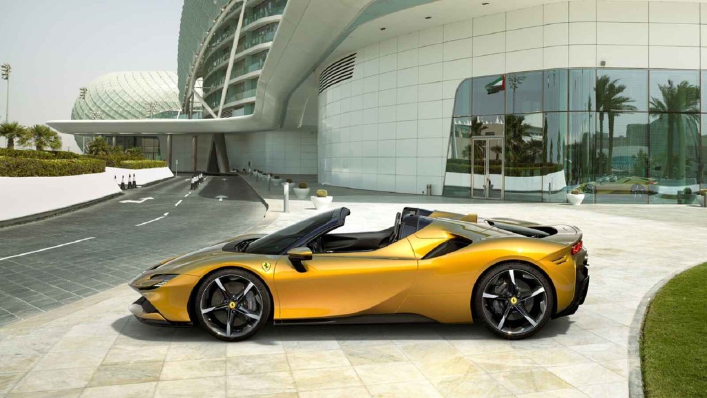 A gold Ferrari SF90 Spider is parked at left side angle with top open in front of a futuristic cement block building