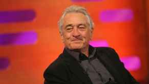 Robert De Niro star of Alto Knights sits smiling in a black suit jacket with a red background
