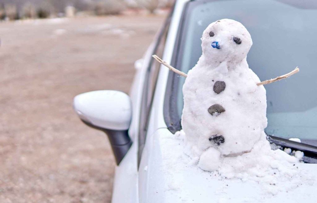 A little snowman is perched on a white car's hood