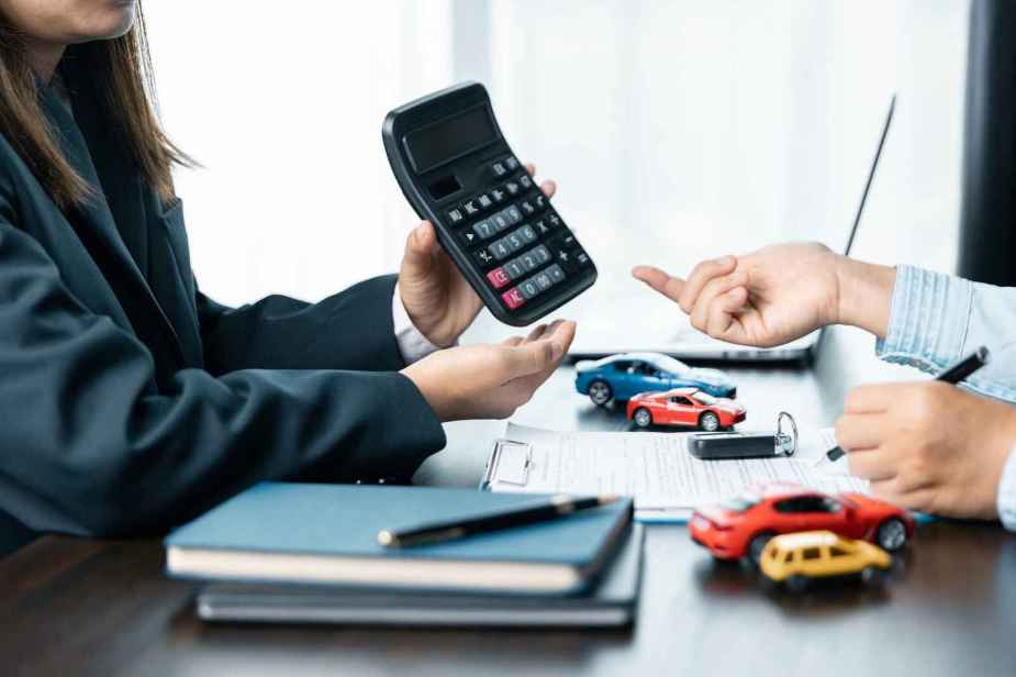 A woman holds a calculator up to a man sitting across from her with journals, toy cars, paperwork on the desk discussing car loan payments or trade-in