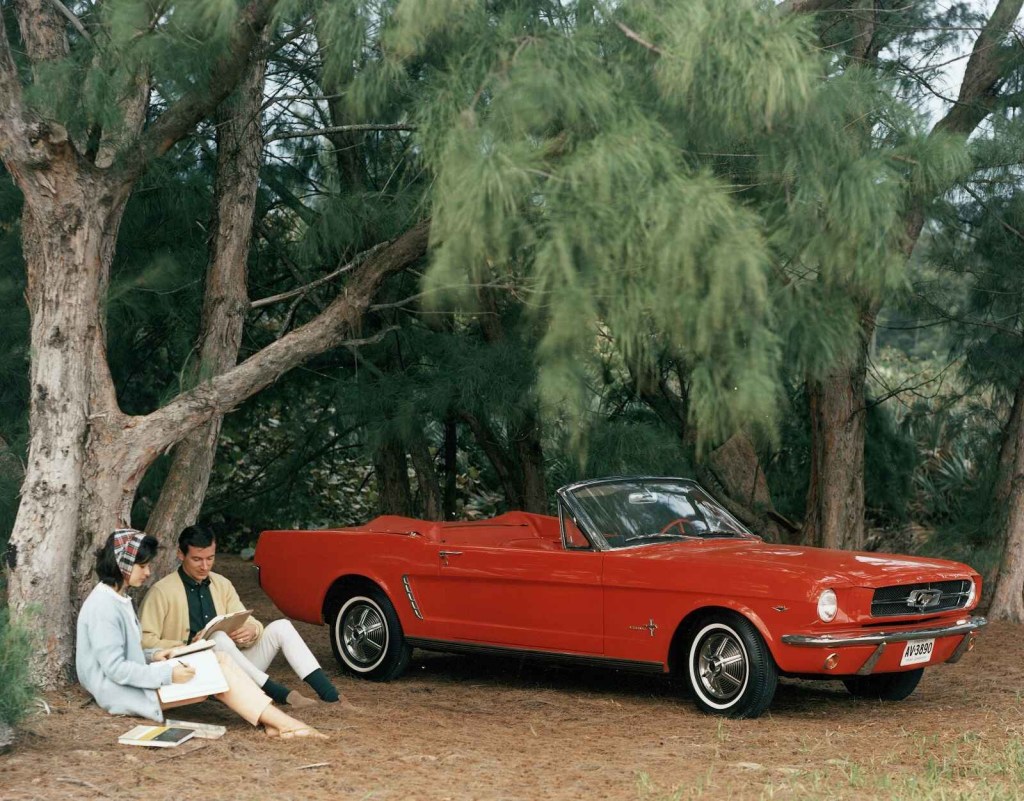 A red 1964 Ford Mustang convertible is parked under a tree with a couple sitting reading on the ground marketing image