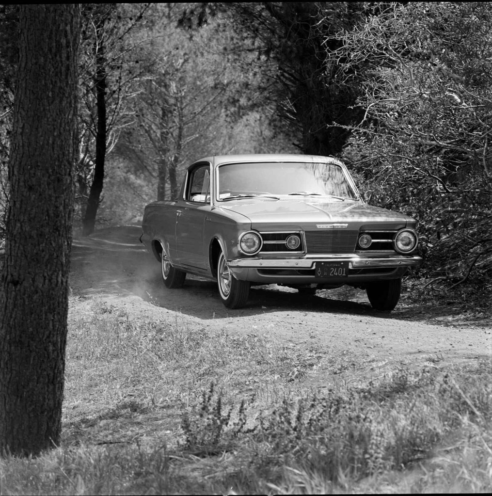 A 1964 Plymouth Barracuda being test driven by MotorTrend though a forested dirt road black and white image
