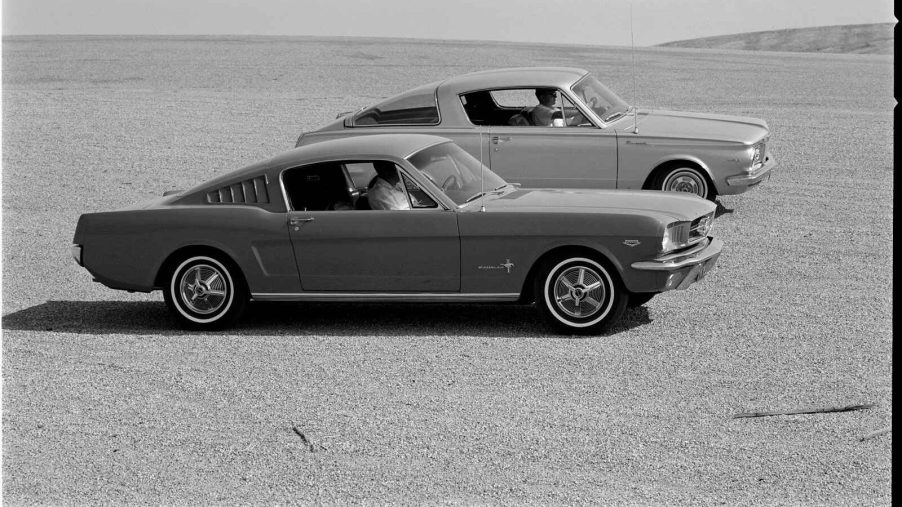 A 1965 Ford Mustang sits next to a 1965 Plymouth Barracuda in the desert the first pony cars black and white photo
