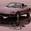 A grey 2004 Mazda MX-5 Miata shown parked on a reflective surface in left front angle view with yellow low beam lights on pink sunset colors