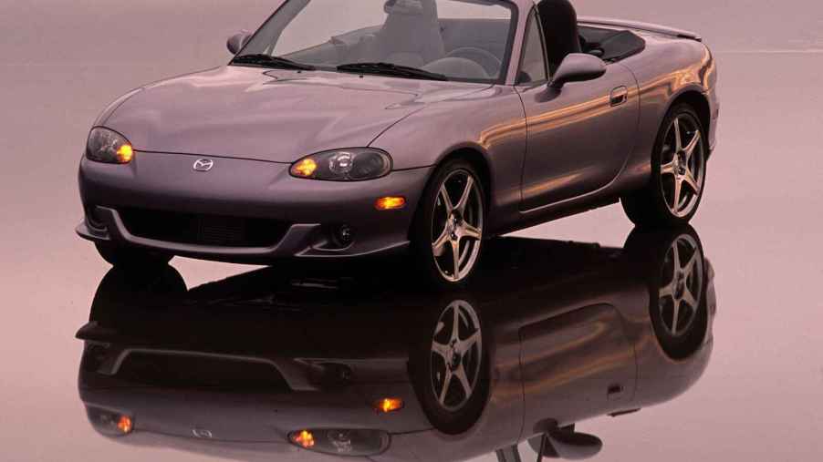 A grey 2004 Mazda MX-5 Miata shown parked on a reflective surface in left front angle view with yellow low beam lights on pink sunset colors