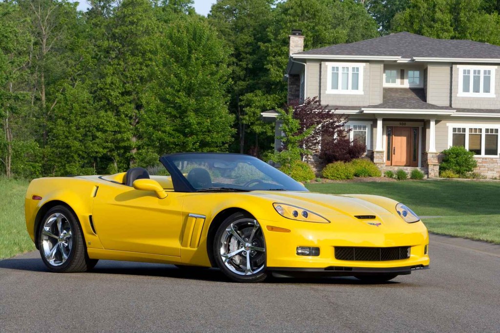 A yellow 2012 Chevrolet Corvette convertible sports car is parked at right front angle view in front of a house and trees